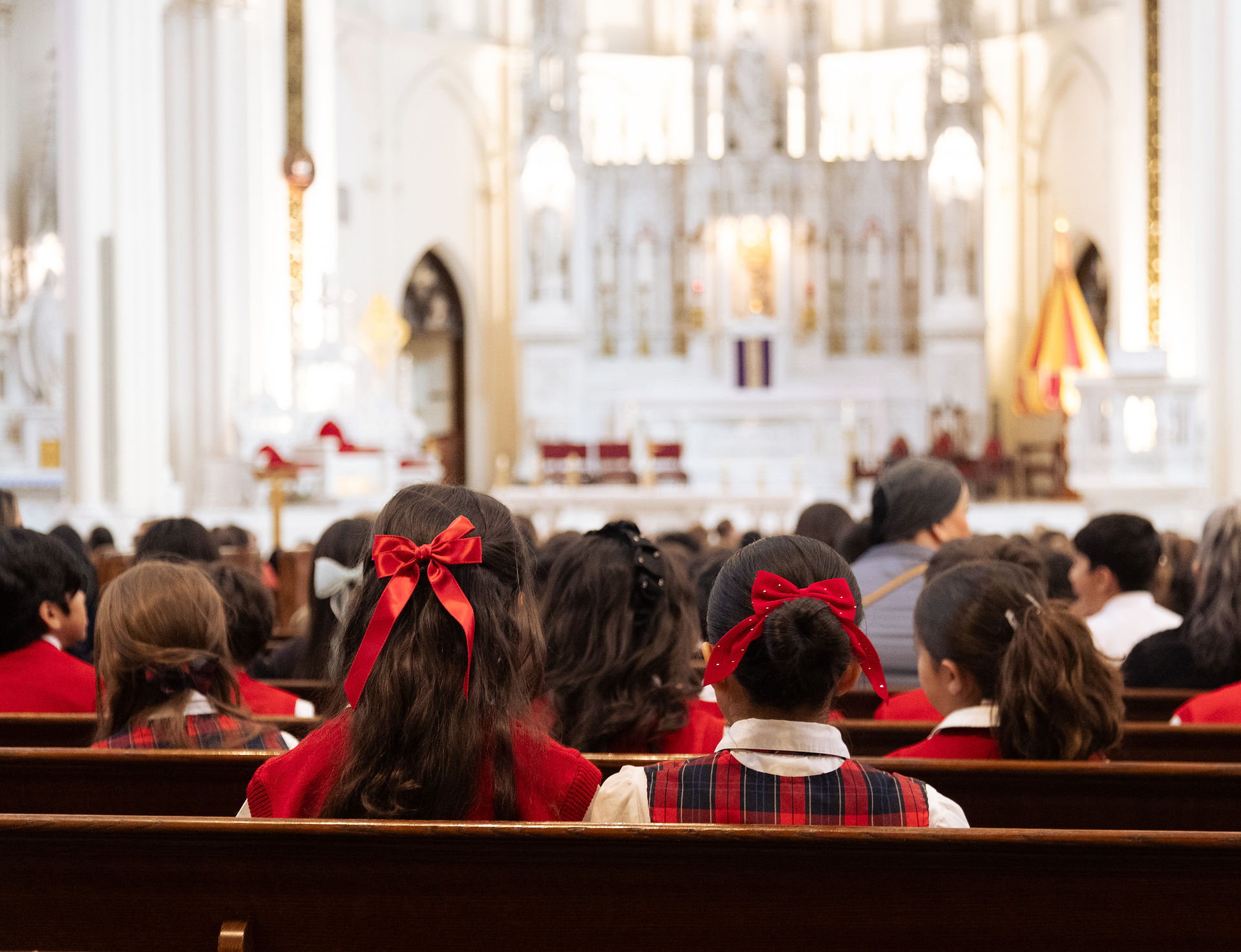 Niñas con lazos rojos en el cabello, sentadas en una iglesia blanca, mirando hacia el altar. Ambiente tranquilo y solemne.