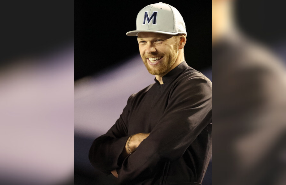 Hombre sonriente con gorra blanca "M", fondo oscuro. Lleva camisa negra y tiene brazos cruzados, aspecto relajado y amigable.