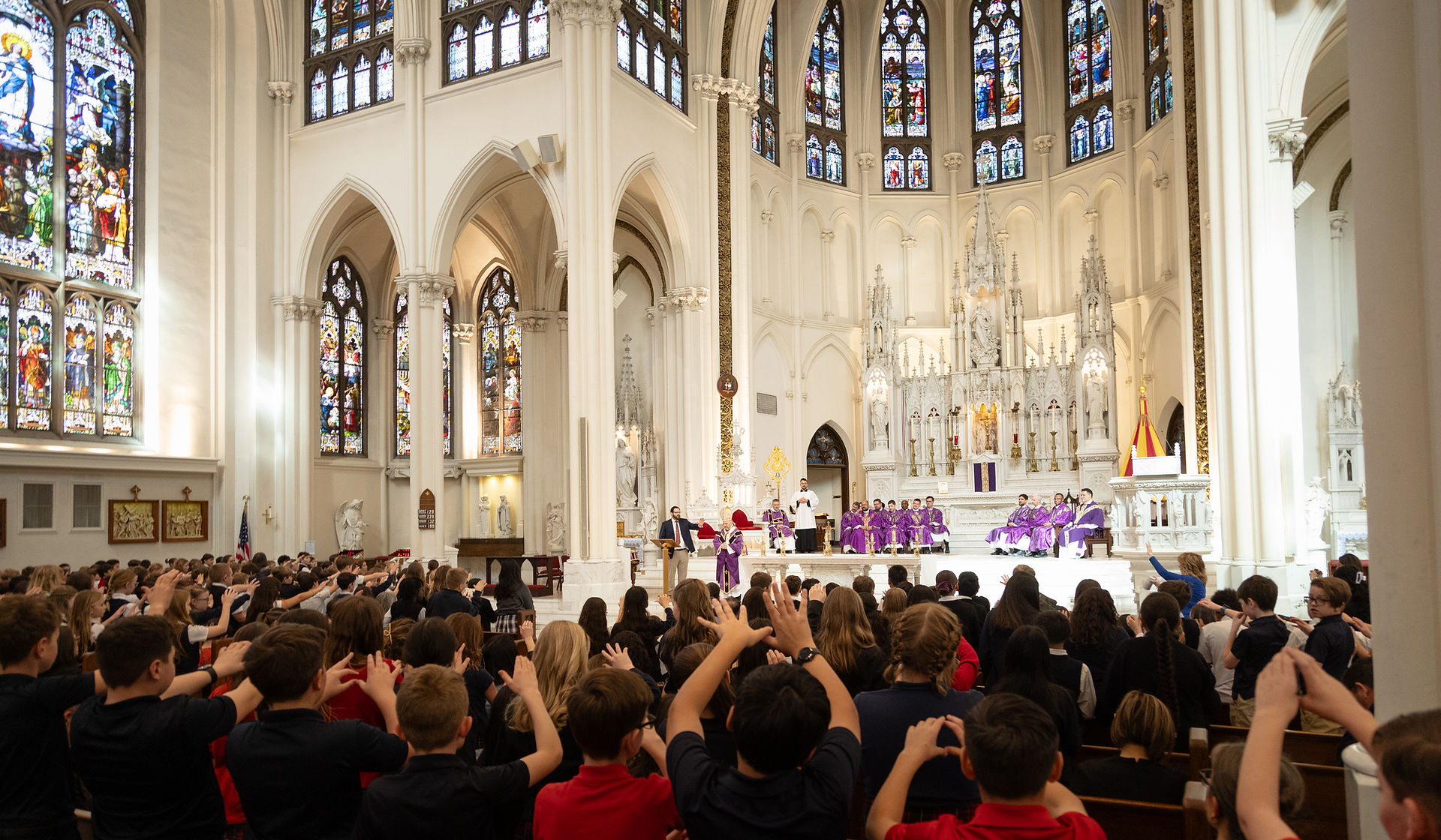 Personas en una iglesia gótica, muchas con manos levantadas hacia el altar donde un sacerdote habla. Vitrales coloridos adornan el fondo.