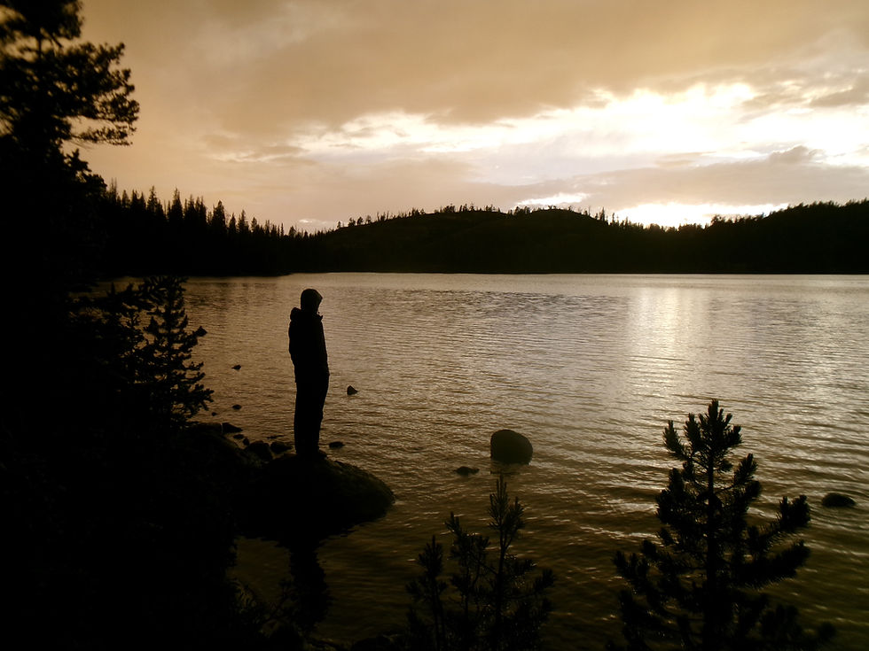 Silueta de una persona junto a un lago al atardecer, con cielo dorado, árboles oscuros y agua tranquila.