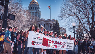 Personas sostienen pancartas "Colorado March for Life" frente a un edificio con cúpula dorada. Ambiente soleado y de protesta pacífica.
