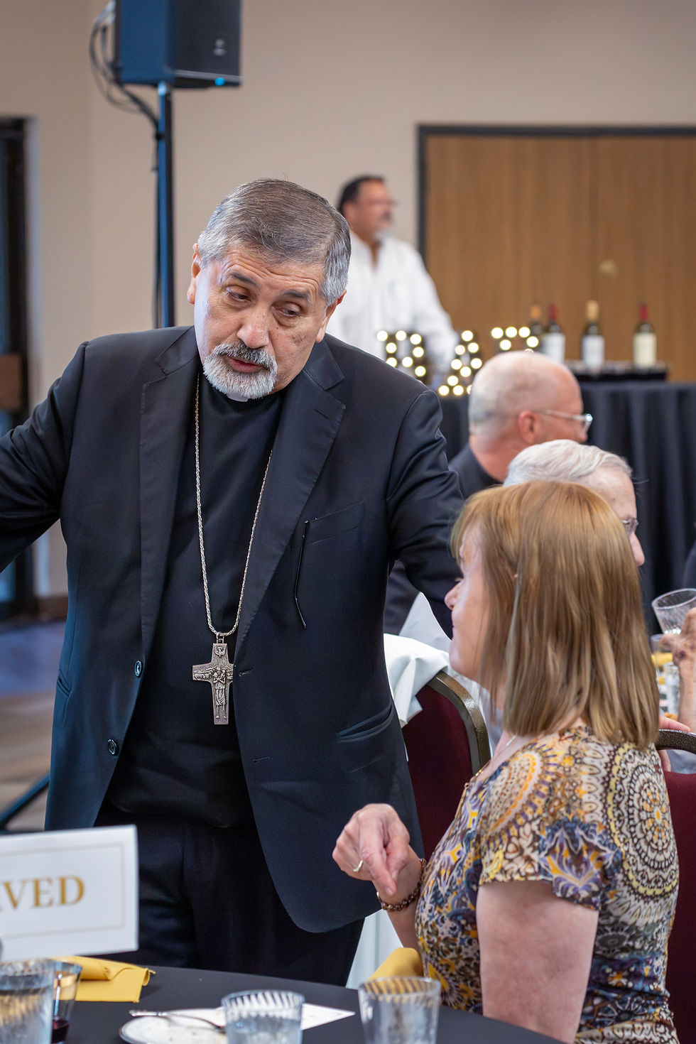 Sacerdote con cruz habla con una mujer sentada en un banquete; fondo desenfocado con invitados y mesas, ambiente serio.