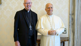 Dos clérigos sonrientes posan juntos: uno en traje negro y otro en blanco, con cruces, ante pared beige decorada.