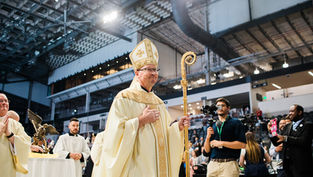 Un obispo en vestimenta dorada sostiene un báculo ceremonial en un evento religioso en un gran salón, rodeado de asistentes y fotógrafos.
