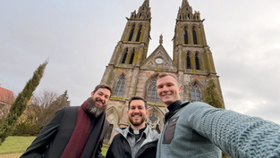 Tres hombres sonrientes posan frente a una iglesia de piedra con dos torres altas. El cielo está nublado, dando un ambiente sereno.