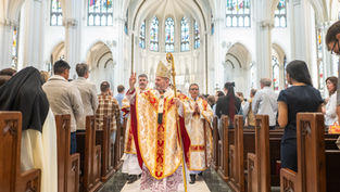 Obispo en túnica dorada y roja bendice a fieles en una catedral con vitrales. Ambiente solemne. Asistentes en bancas observan atentos.