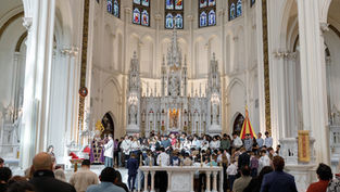 Personas en una iglesia gótica, frente al altar blanco, participan en una ceremonia. Luz suave entra por las vidrieras coloridas.
