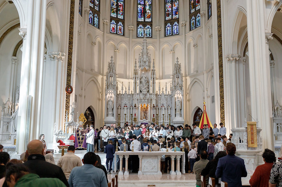 Personas en una iglesia gótica, frente al altar blanco, participan en una ceremonia. Luz suave entra por las vidrieras coloridas.