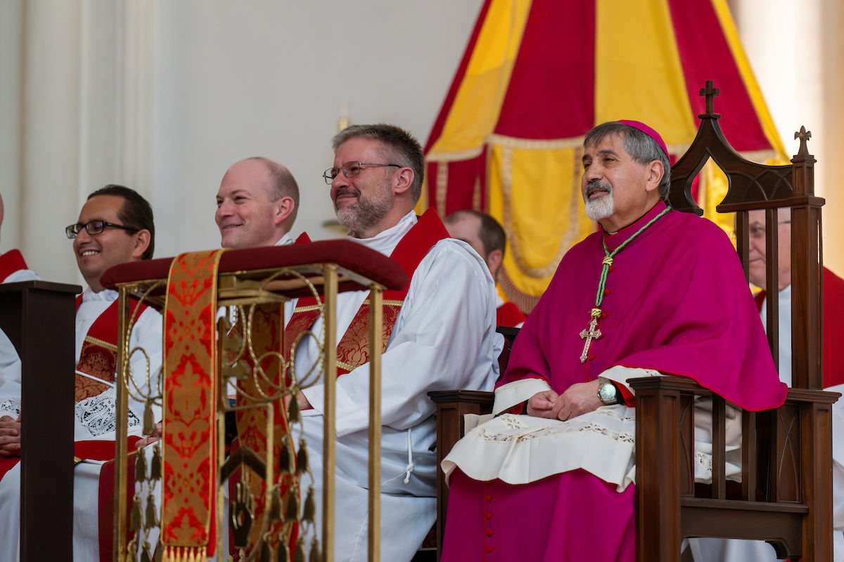 Clérigos sentados cerca del santuario, incluido un obispo con vestiduras rojas, escuchan atentamente durante la liturgia.