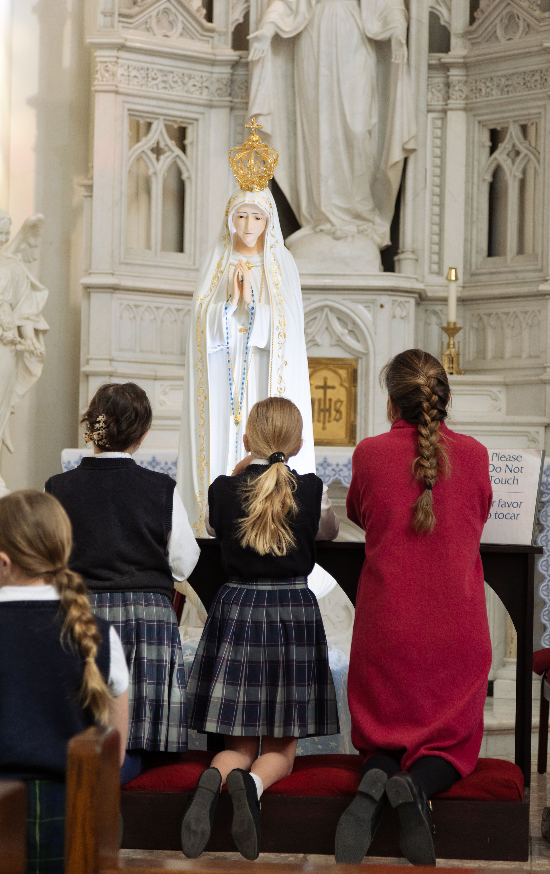Niñas y una mujer rezan arrodilladas ante una estatua de la Virgen en una iglesia. Visten uniformes oscuros y el fondo es de mármol detallado.