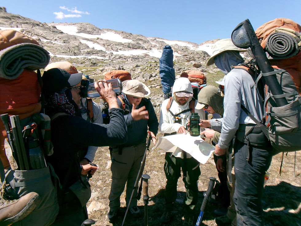 Grupo de excursionistas con mochilas y bastones consulta un mapa en un paisaje rocoso de montaña con restos de nieve y cielo despejado.