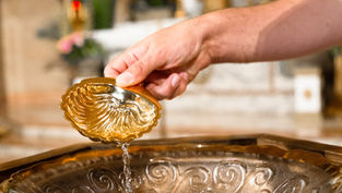 Mano sostiene una copa dorada en forma de concha y vierte líquido en un cuenco ornamentado; fondo desenfocado.