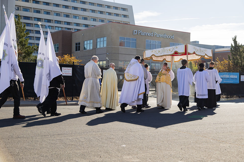Procesión religiosa frente a un edificio de Planned Parenthood. Participantes con túnicas y capas blancas portan estandartes y un dosel decorado. Ambiente solemne.