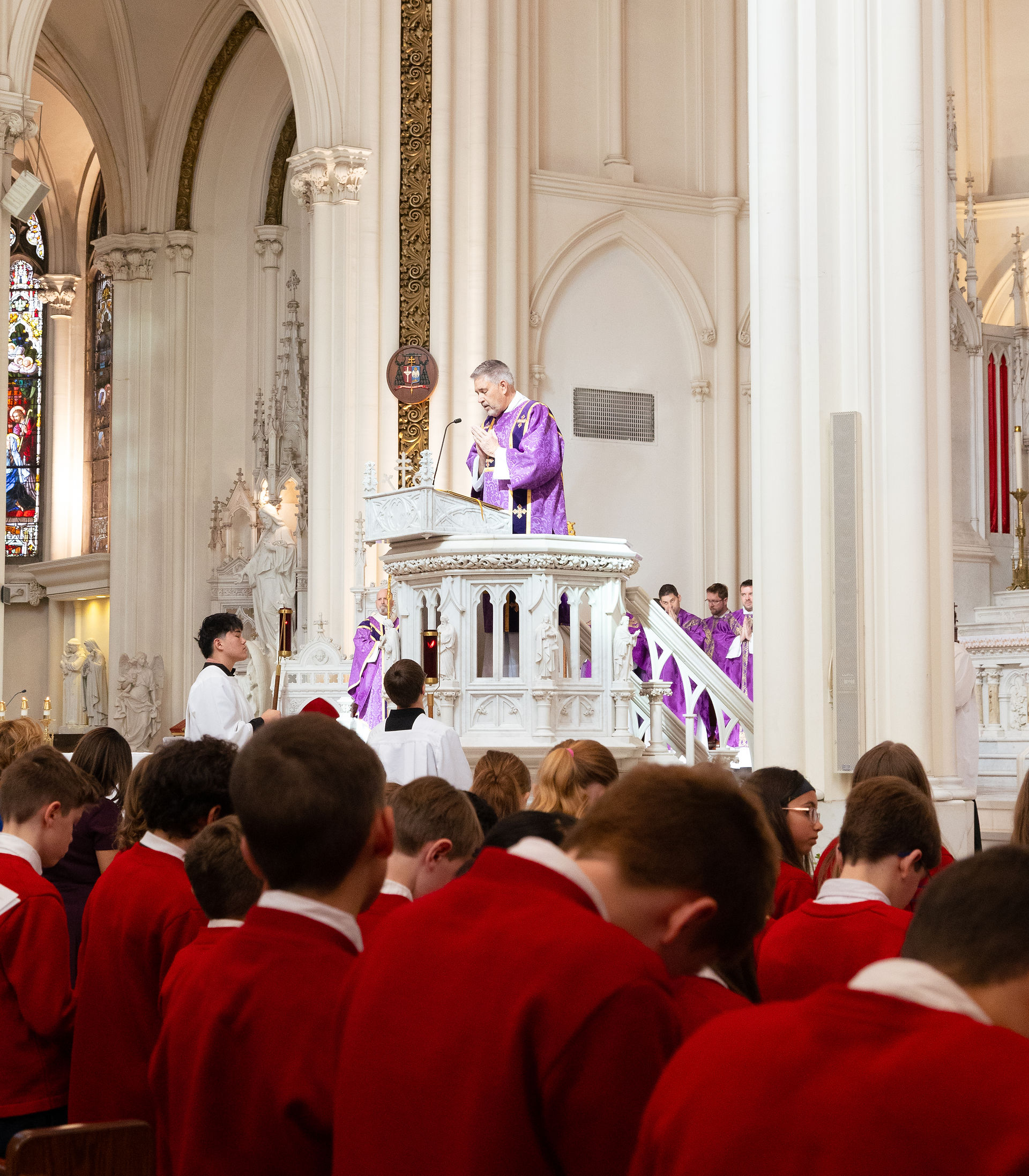 Diácono en púlpito con vestimenta púrpura en una iglesia, hablando ante una congregación de niños y jóvenes en ropas rojas. Ambiente solemne.