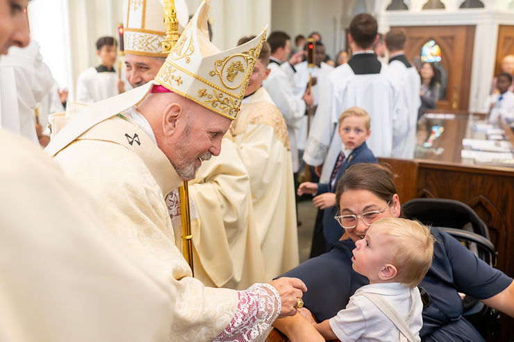 Obispo con vestiduras doradas y mitra sonríe y saluda a un niño pequeño en brazos de su madre durante una liturgia en la iglesia, con clero y feligreses al fondo.