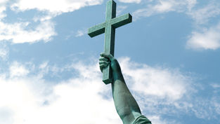 Brazo de estatua verde sosteniendo una cruz de madera contra un cielo azul con nubes, evocando solemnidad.