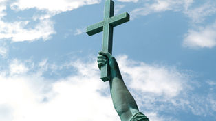 Brazo de estatua verde sosteniendo una cruz de madera contra un cielo azul con nubes, evocando solemnidad.