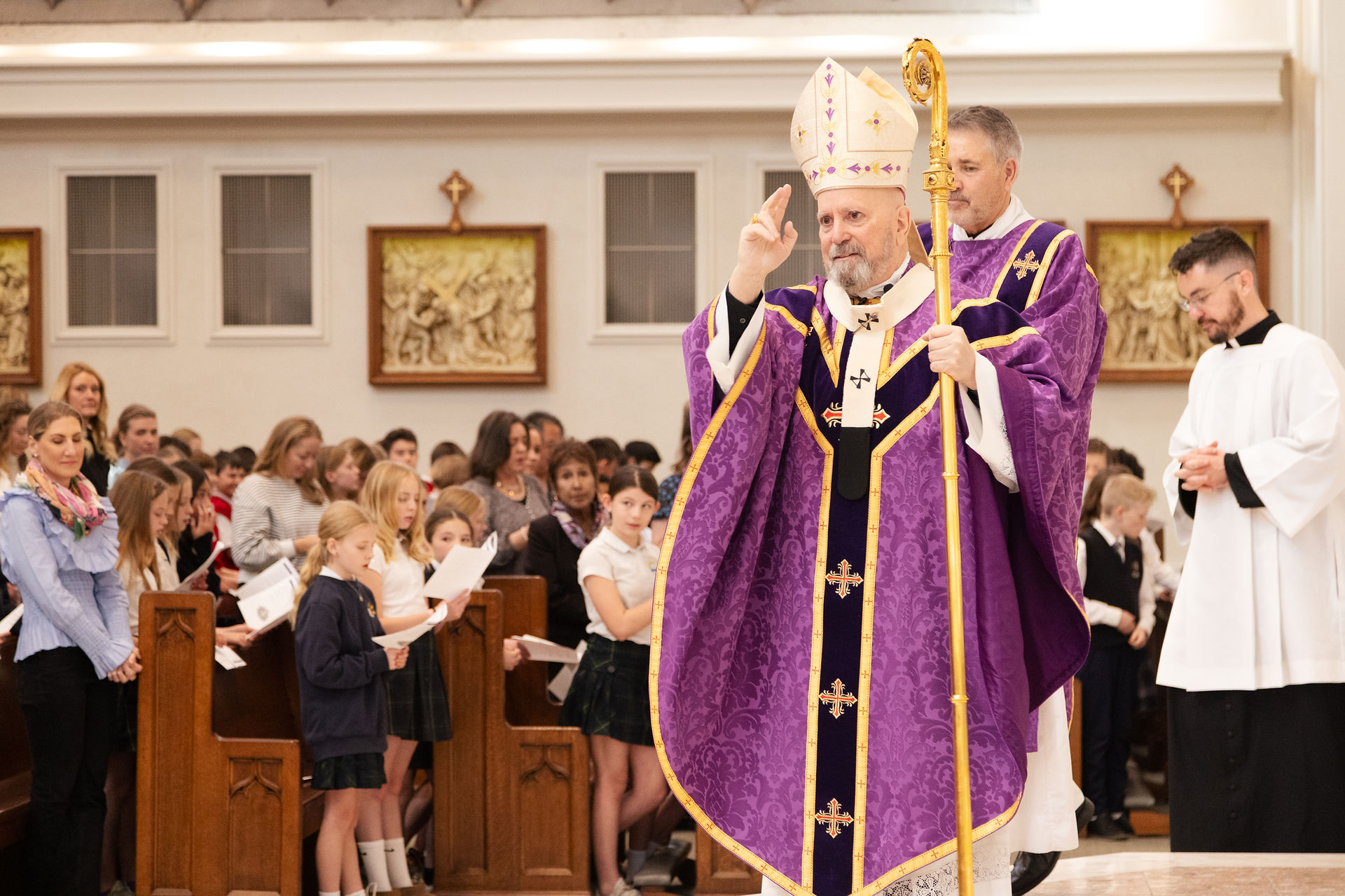 Obispo con vestiduras moradas y mitra levanta la mano en señal de bendición mientras camina por el pasillo de una iglesia llena de estudiantes y feligreses.