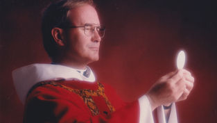 Sacerdote con túnica roja y blanca sostiene una hostia iluminada sobre fondo rojo; escena solemne y contemplativa.