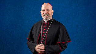 Un hombre con atuendo religioso negro y detalles rojos, sonriendo frente a un fondo azul. Lleva un anillo y collar. Ambiente formal.