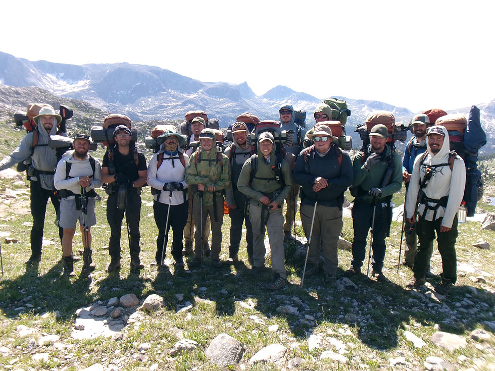 Grupo de excursionistas con mochilas y bastones posa en una montaña rocosa, con picos nevados al fondo y sonrisas.