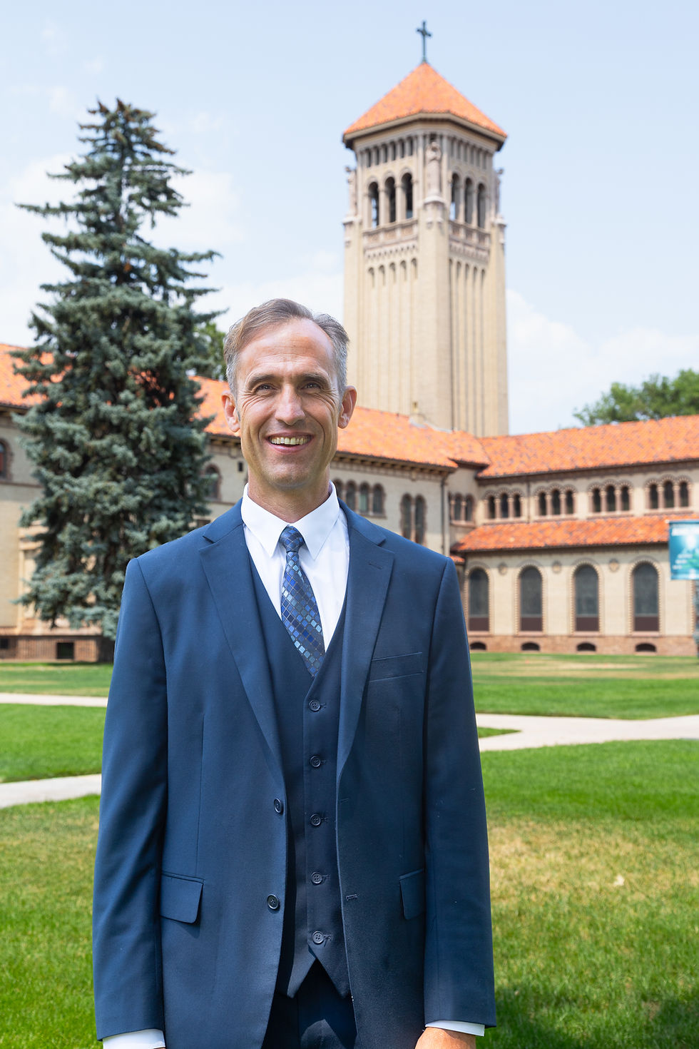 Hombre sonriente con traje azul frente a una torre de iglesia con tejado naranja y césped en un campus soleado.