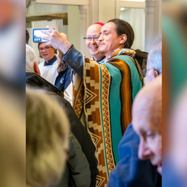Hombre con poncho colorido se toma una selfie sonriente entre una multitud en interiores.
