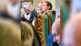 Hombre con poncho colorido se toma una selfie sonriente entre una multitud en interiores.