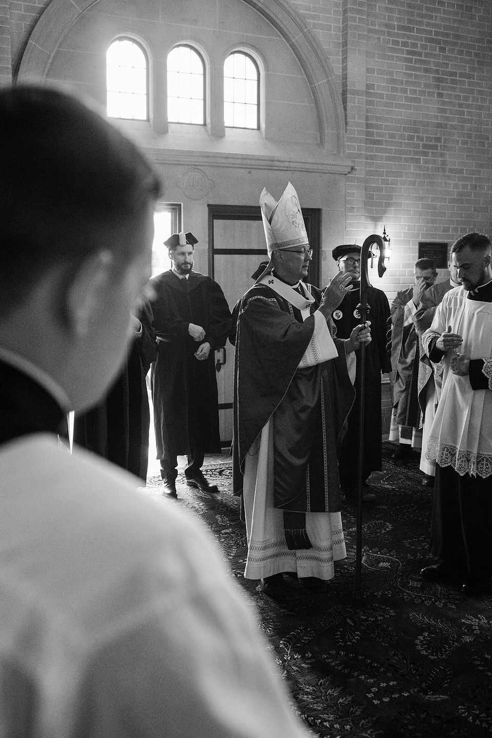 Black-and-white church ceremony with a bishop in mitre blessing clergy inside a brick sanctuary with arched windows.