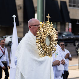 Sacerdote con custodia dorada en una procesión religiosa urbana, rodeado de fieles con vestiduras blancas y banderas.