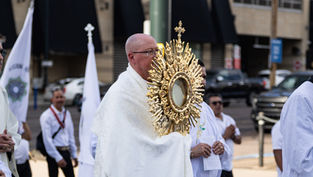 Sacerdote con custodia dorada en una procesión religiosa urbana, rodeado de fieles con vestiduras blancas y banderas.