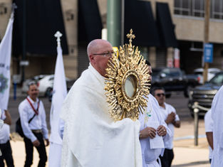Sacerdote con custodia dorada en una procesión religiosa urbana, rodeado de fieles con vestiduras blancas y banderas.