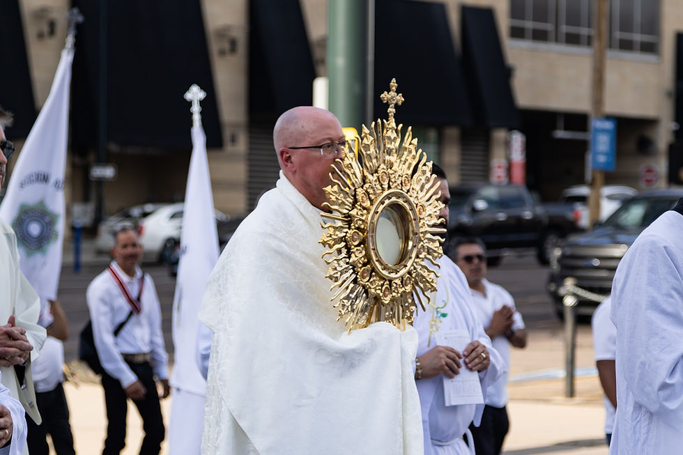 Sacerdote con custodia dorada en una procesión religiosa urbana, rodeado de fieles con vestiduras blancas y banderas.