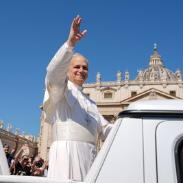 El papa sonriente saluda desde un vehículo blanco en la Plaza de San Pedro, con la basílica al fondo y cielo azul.