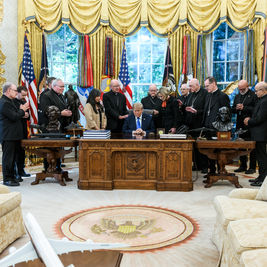 Trump sentado en el Despacho Oval, rodeado de clérigos en oración, con banderas y cortinas doradas de fondo.