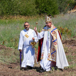 Dos clérigos con vestiduras blancas celebran una ceremonia al aire libre; uno alza un báculo y el otro sostiene un cáliz.