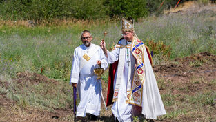 Dos clérigos con vestiduras blancas celebran una ceremonia al aire libre; uno alza un báculo y el otro sostiene un cáliz.