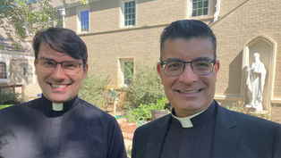 Dos sacerdotes sonrientes con gafas posan ante un edificio de ladrillo y una estatua blanca en un patio soleado.