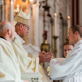 Tres clérigos en una iglesia se dan la mano durante una ceremonia, con vestimentas blancas y mitra beige; ambiente solemne.