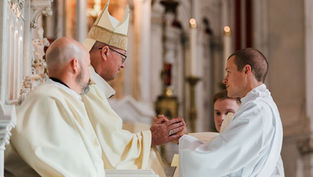 Tres clérigos en una iglesia se dan la mano durante una ceremonia, con vestimentas blancas y mitra beige; ambiente solemne.