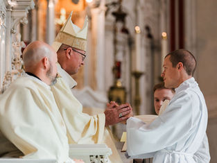 Tres clérigos en una iglesia se dan la mano durante una ceremonia, con vestimentas blancas y mitra beige; ambiente solemne.