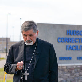 Sacerdote de negro con cruz, gesto solemne, frente al letrero de Hudson Correctional Facility en Colorado.