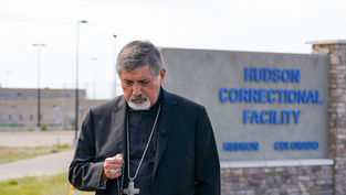 Sacerdote de negro con cruz, gesto solemne, frente al letrero de Hudson Correctional Facility en Colorado.