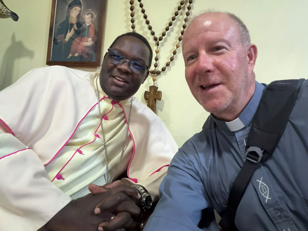 Dos sacerdotes sonrientes se toman una selfie en una habitación con rosario grande y cuadro religioso.