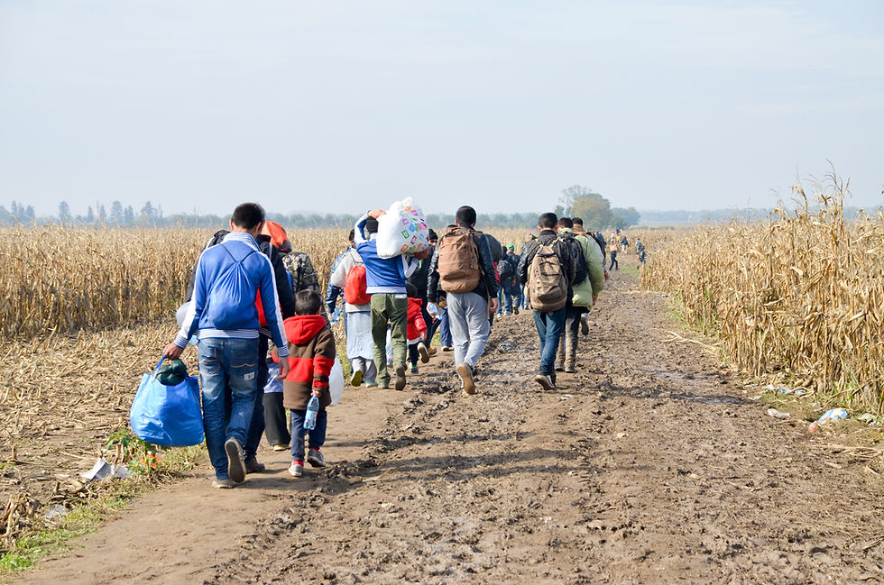 Grupo de personas y niños camina con mochilas y bolsas por un sendero lodoso entre maizales secos bajo cielo despejado.