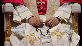 Pope con vestiduras blancas y cruz dorada, sentado, sosteniendo un rosario negro; detalles bordados en oro.