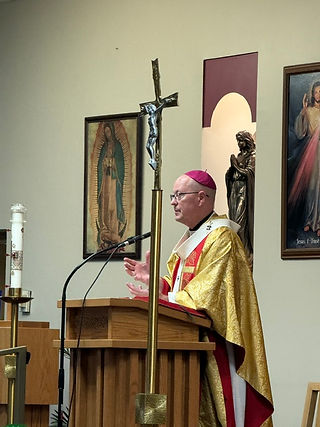 Obispo con capa dorada y solideo rosa habla desde el púlpito en una iglesia, junto a un crucifijo y cuadros religiosos.