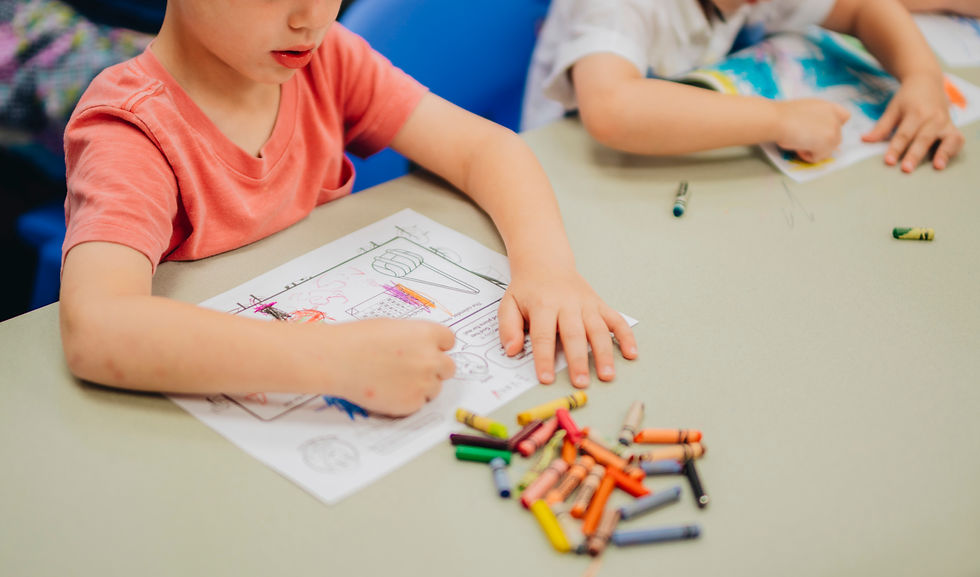Niños coloreando dibujos en una mesa con crayones dispersos; uno lleva camiseta rosa y el ambiente es de clase tranquila.