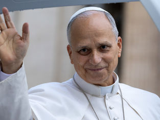 El papa León sonríe y saluda con la mano, vestido de blanco, en un fondo interior desenfocado.