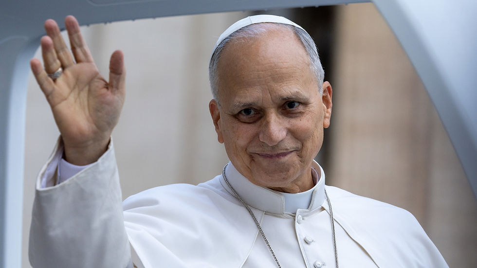 El papa León sonríe y saluda con la mano, vestido de blanco, en un fondo interior desenfocado.
